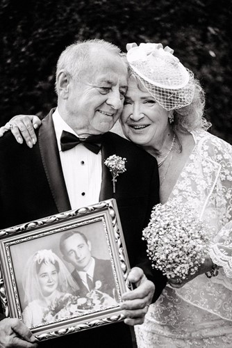 Older couple dressed for wedding holding picture of them as younger bride and groom